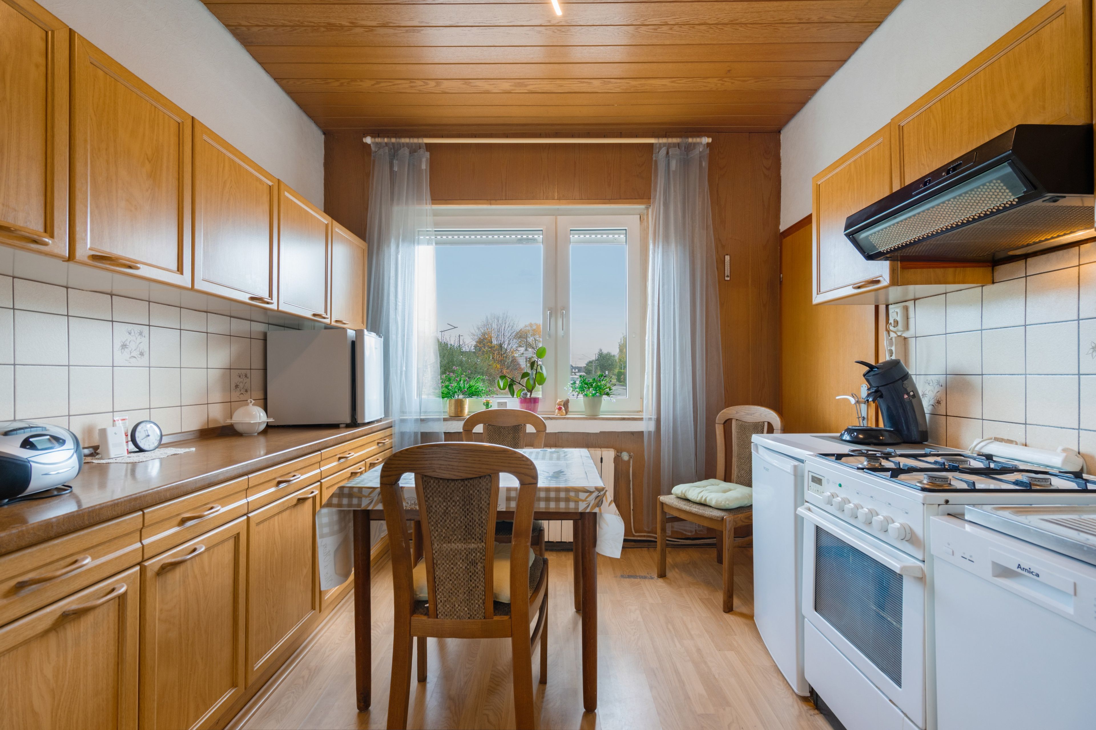 A kitchen with wooden cabinets, a dining table with chairs, a window with curtains, and a stove.