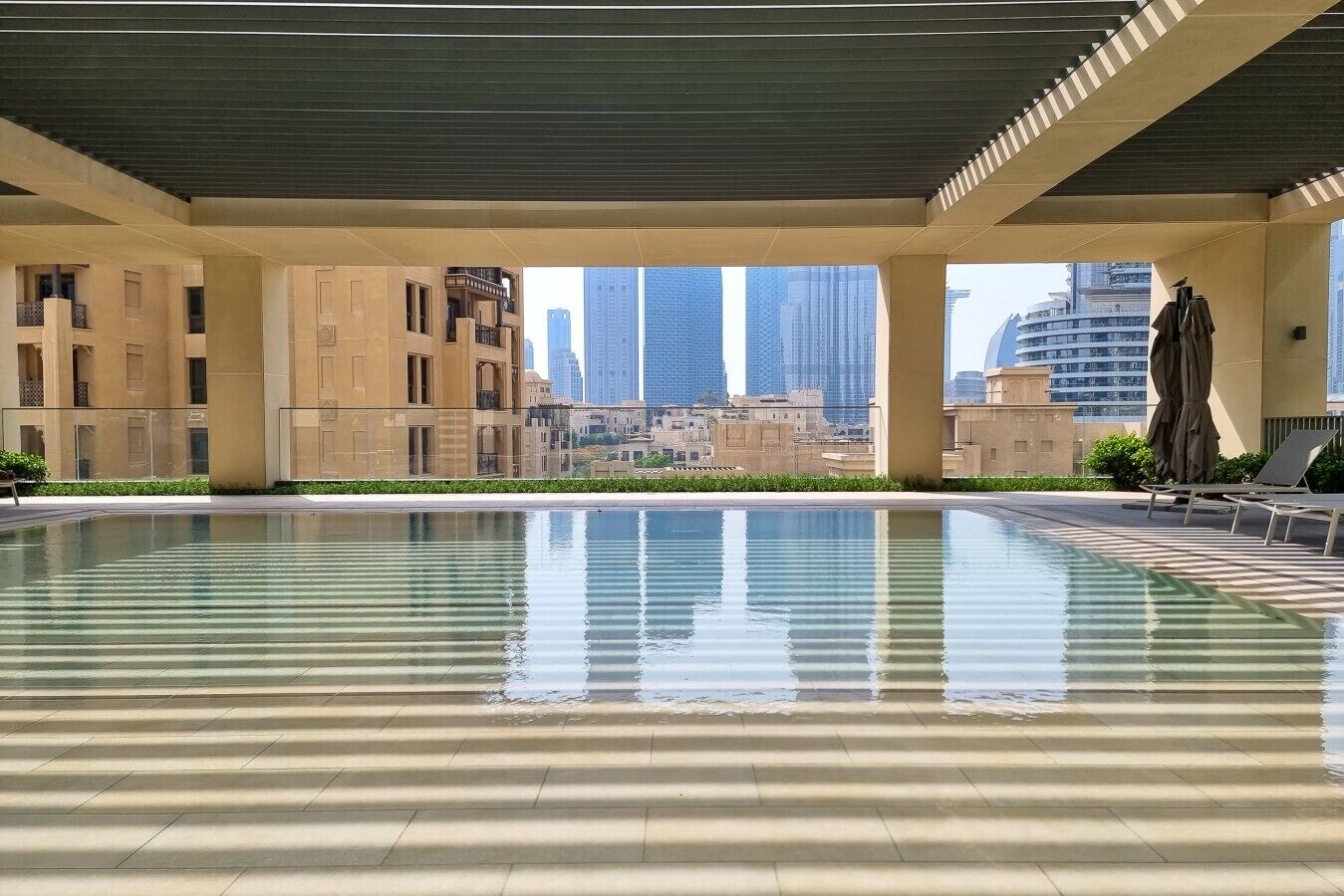 Une piscine accessible par des marches, entourée d’une zone abritée d’où l’on peut admirer de grands immeubles.