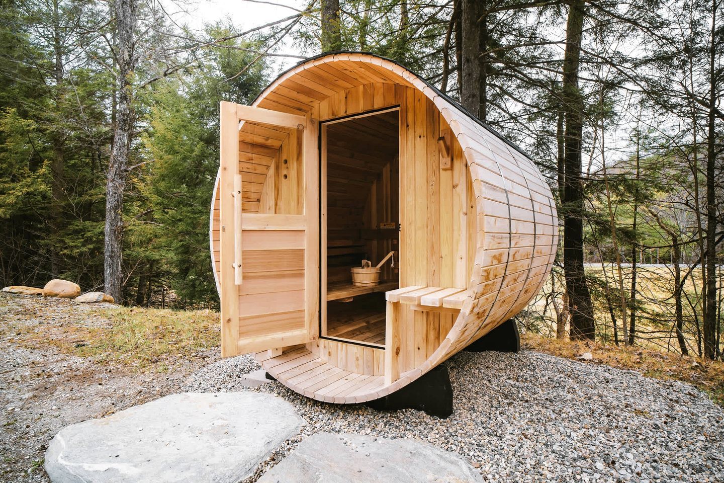 A wooden sauna with a curved roof, open door, and benches inside, set in a forest.