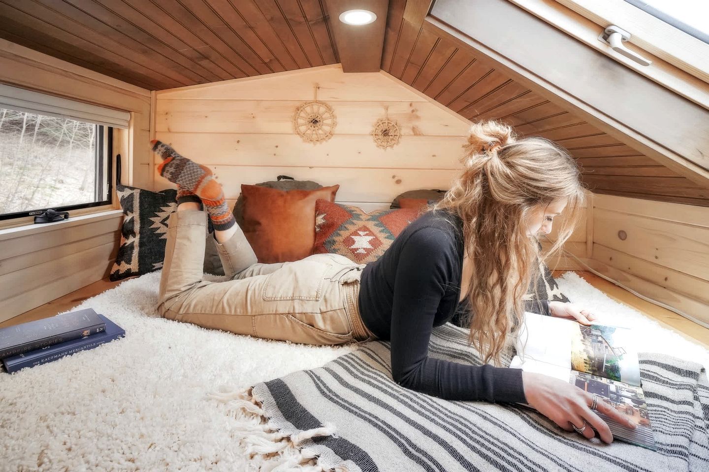 A cozy attic room with a sloped ceiling, a window with blinds, a patterned rug, and a person lying on the floor.