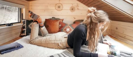 A cozy attic room with a sloped ceiling, a window with blinds, a patterned rug, and a person lying on the floor.