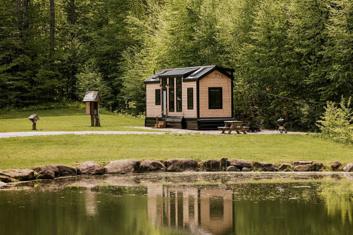 A cabin with a small porch, a picnic table, and a grill, surrounded by a pond and dense forest.