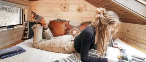 A cozy attic room with a sloped ceiling, a window with blinds, a patterned rug, and a person lying on the floor.