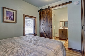 Une chambre à coucher avec un lit, une fenêtre avec des stores, une porte et une salle de bain visible à travers une porte ouverte.