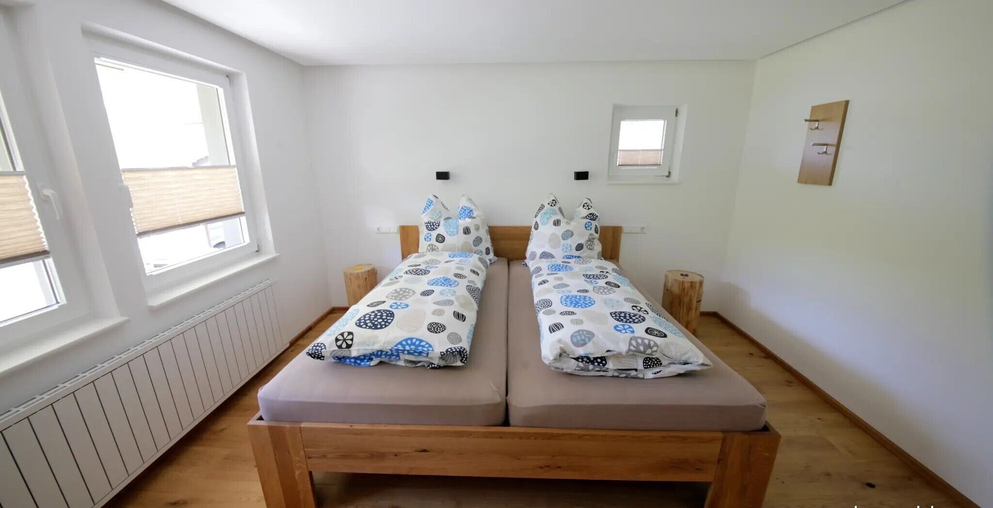 A twin bed with patterned bedding, a wooden headboard, a small bedside table, a radiator, and a window with blinds.