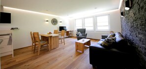 A living room with a wooden floor, a black sofa, a dining table with chairs, a TV, and a clock on the wall. - Apartment 'Abendgrund' with Private Terrace, Shared Garden and Wi-Fi (Unterkirnach)