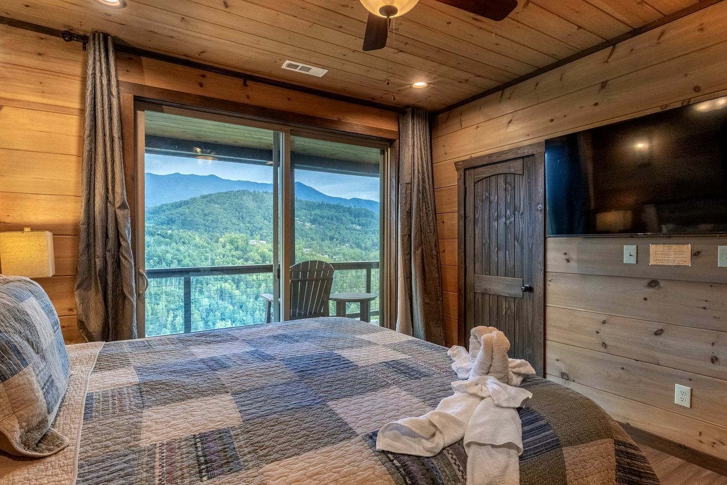 Ein Schlafzimmer in einer Holzhütte mit einem großen Bett, einem Sessel, einem Fernseher und einem Balkon mit Blick auf die Berge.