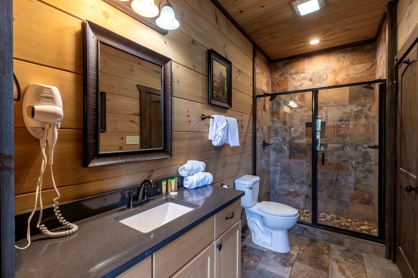 Une salle de bain avec un meuble-lavabo en bois, un évier, des toilettes, une douche aux murs en pierre et un sèche-cheveux.