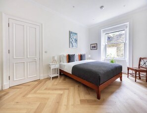A bedroom with a wooden floor, a bed with a dark blanket, a nightstand, a chair, a window with a view, and a door. - Spacious 1BR in Heart of New Town & Royal Crescent (Edinburgh)