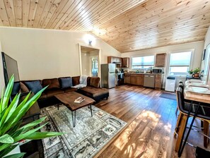 A spacious living area with a sofa, coffee table, and a rug. A kitchen with wooden cabinets and a dining area with chairs is visible. - Peaceful Garden Retreat Near Acadia National Park, Maine (Ellsworth)