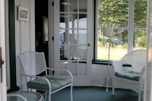 A room with a door leading outside, two wicker chairs with cushions, a small table, and a picture on the wall. - Scenic Waterfront Summer Cottage with Ocean Views, Kayaking & Beach Access near Bar Harbor, Hancock Point, Maine (Hancock)