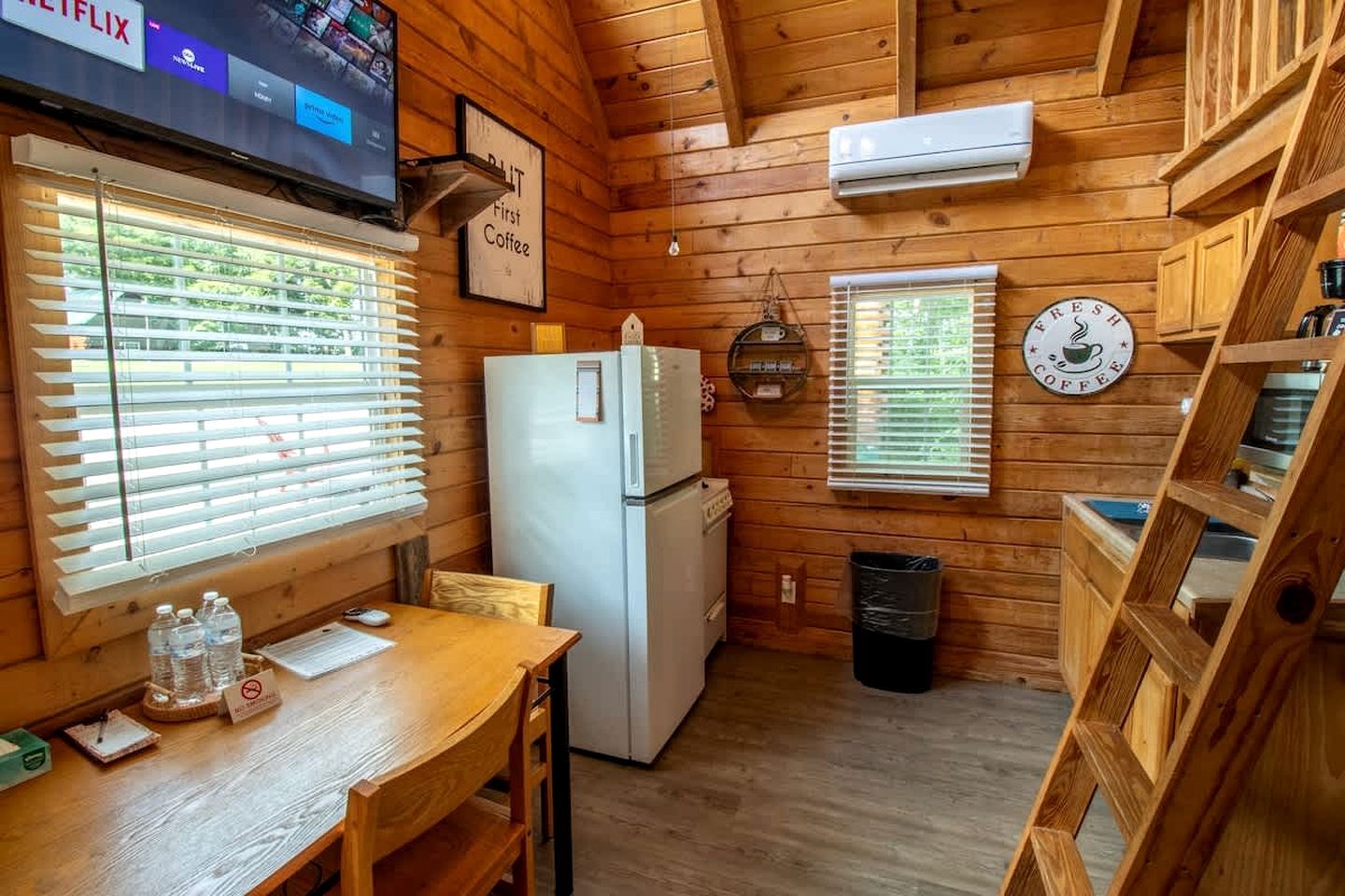 A wooden cabin interior with a kitchen area, a dining table, a refrigerator, a TV mounted on the wall, and a sign indicating 'First Coffee.'