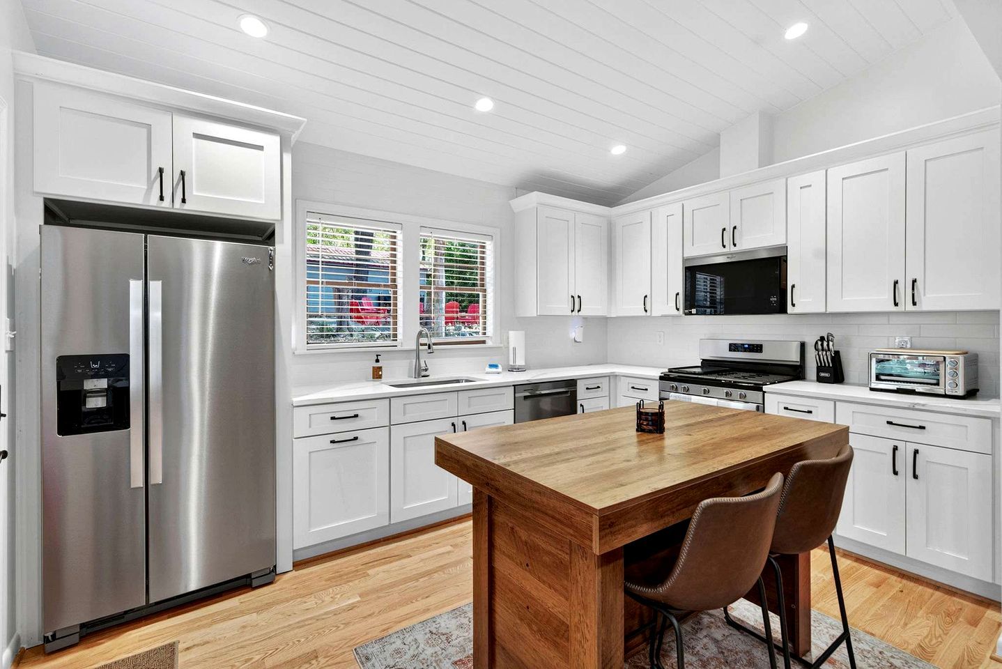 A modern kitchen with white cabinets, stainless steel appliances, a wooden island, and a view of the outside through large windows.