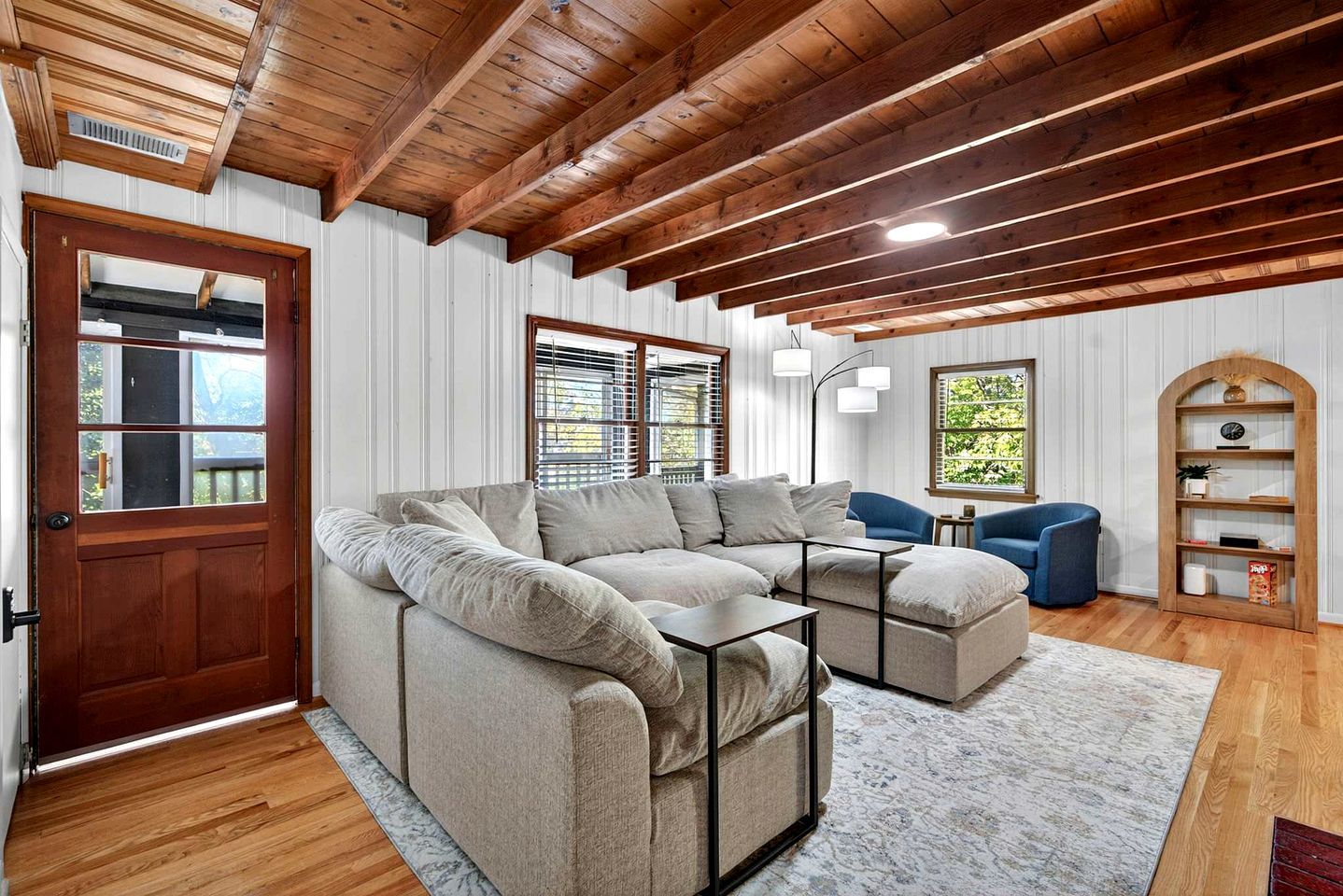 A cozy living room with a sectional sofa, a blue armchair, a wooden bookshelf, and a door with a window.