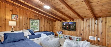 Una habitación tipo cabaña con paredes y techo de madera, una cama con ropa de cama azul, un pequeño sofá, un sillón de peluche y un televisor montado en la pared, además de una ventana con persianas.