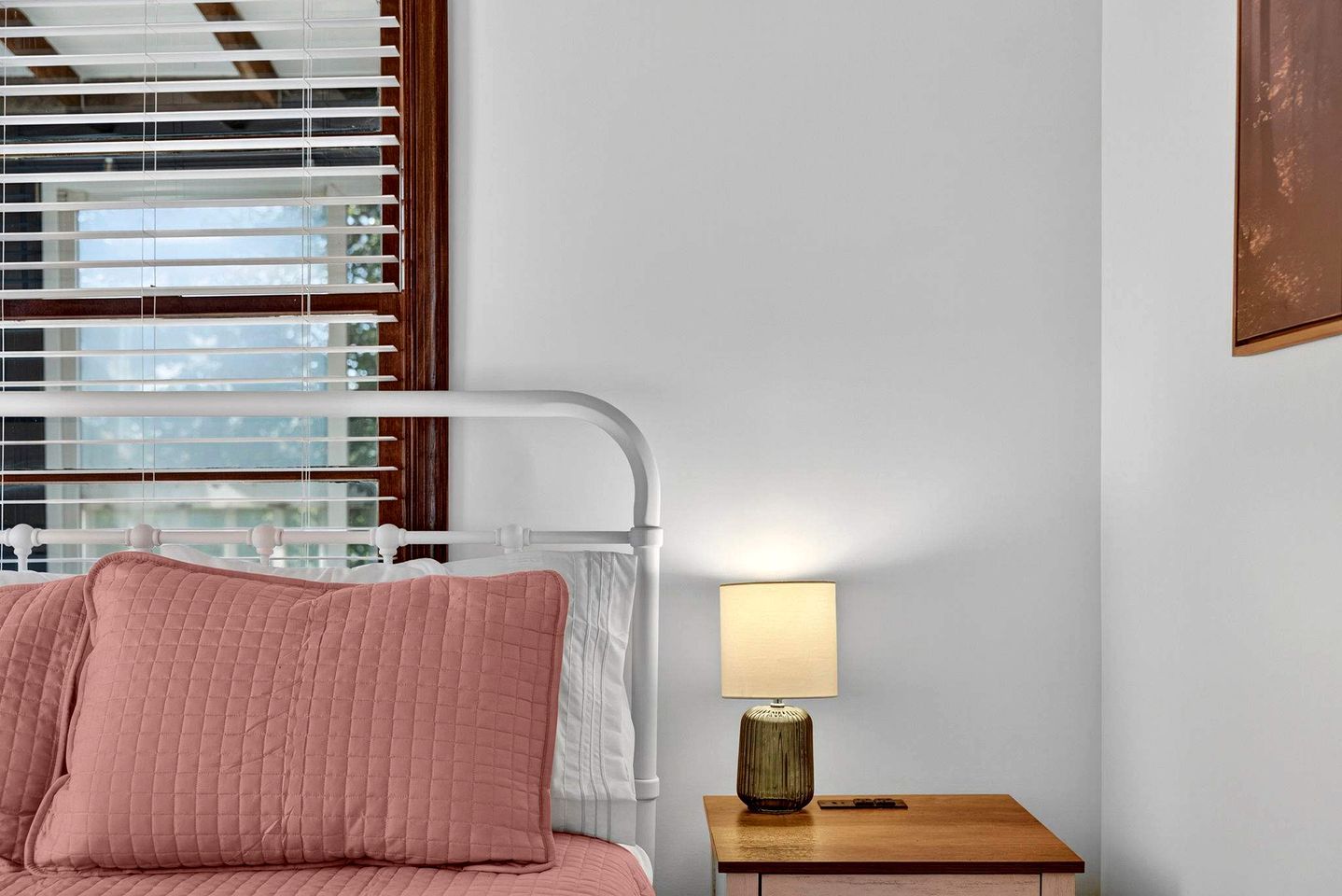 Een slaapkamer met een bed, een bedlamp, een nachtkastje en een raam met jaloezieën.