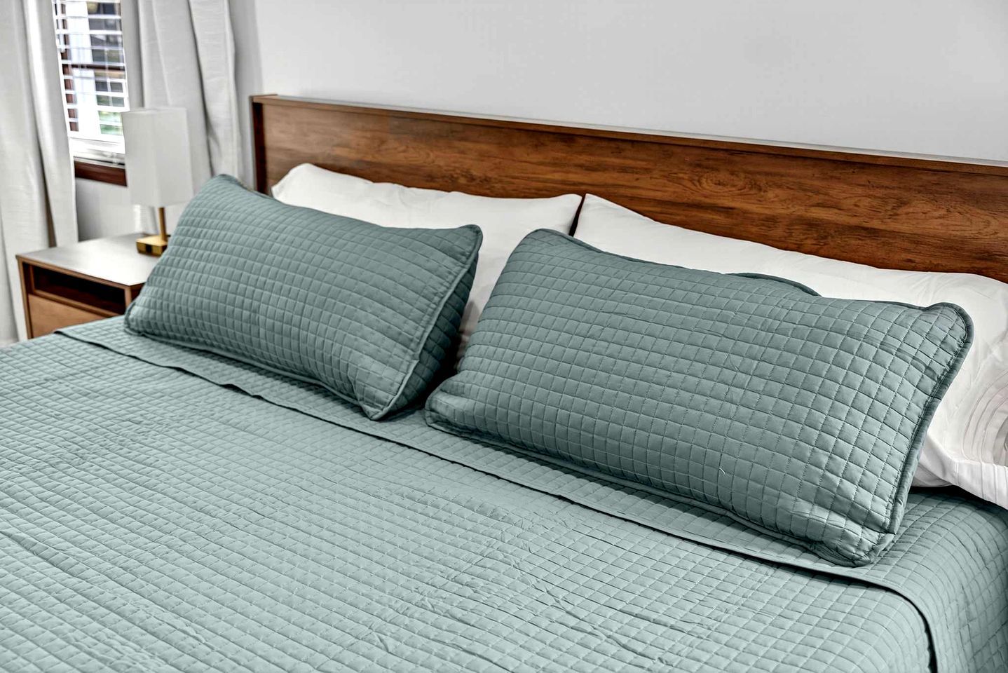 Una cama bien hecha con colcha verde acolchada y almohadas, un cabecero de madera y una mesita de noche con lámpara.
