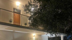 Un edificio de dos pisos con balcón, una banca, plantas en macetas y piso de baldosas.
