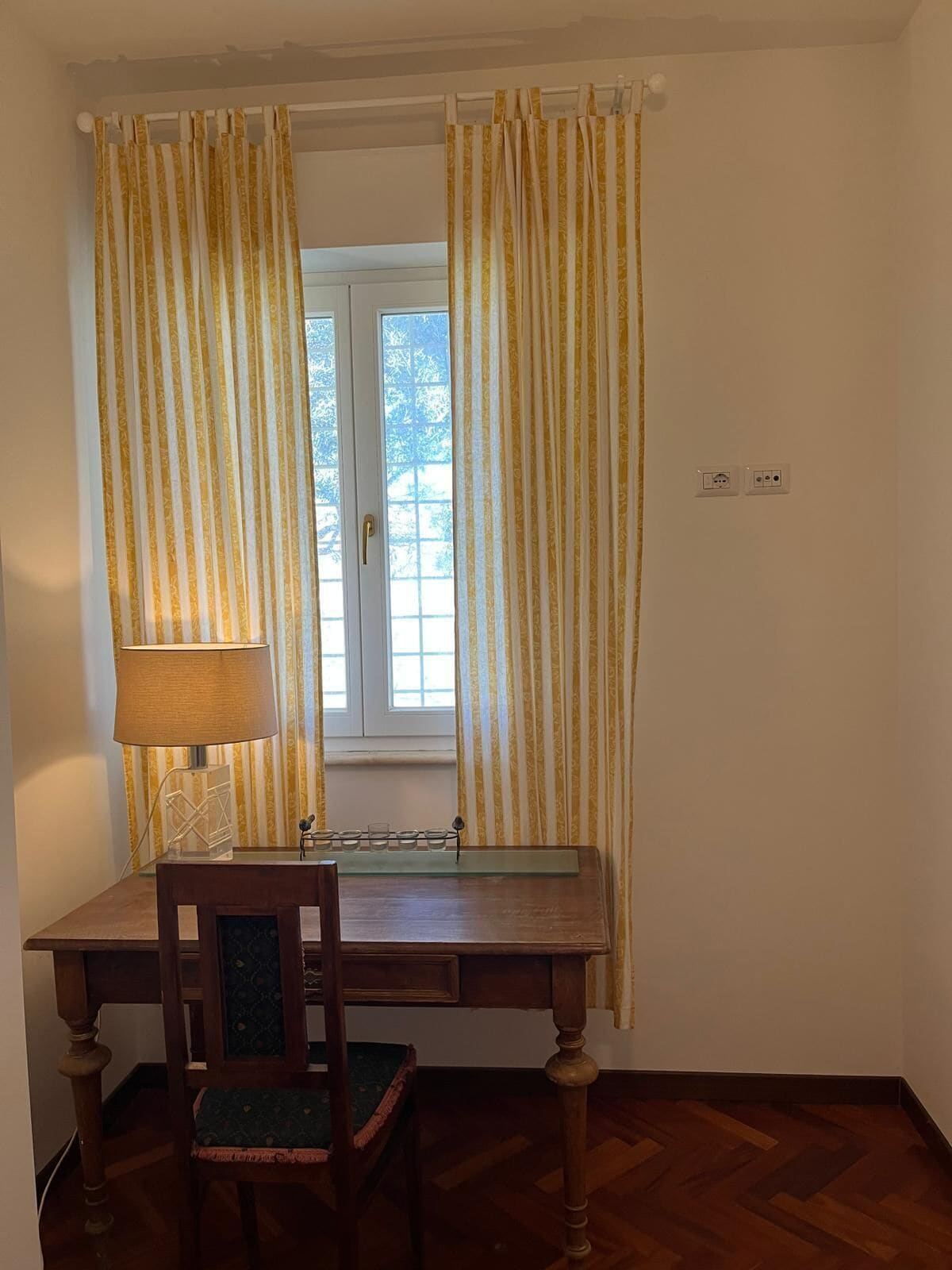 Una habitación con un escritorio de madera, una silla, una lámpara y una ventana con cortinas.