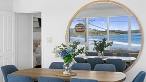 A dining area with a large round mirror reflecting a coastal view, a wooden table, and blue upholstered chairs.