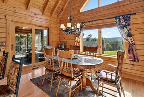 Interior de una cabaña de madera con mesa de comedor, sillas, una araña y ventanas amplias que ofrecen vistas al exterior.