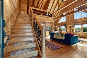 A wooden staircase with a decorative railing, a blue sofa, and a large window in a cabin-style interior. - Peaceful Mountain Cabin with Hot Tub, Screened Porch & Views Near Lost River, Baker WV (Baker)