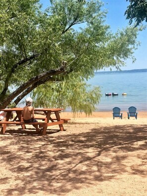 Une table de pique-nique avec deux chaises Adirondack, placée sous un arbre, près d’un lac.