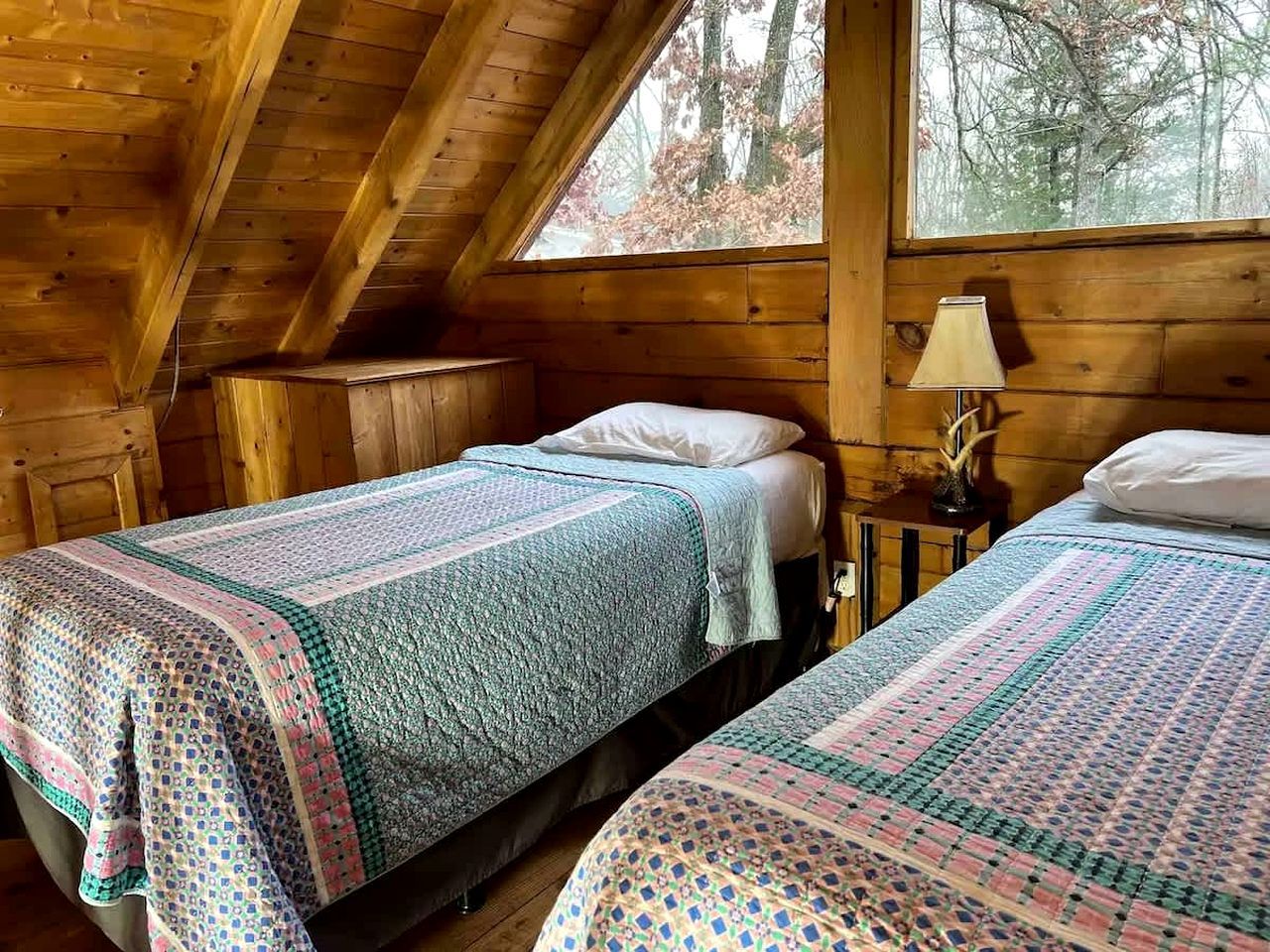 A cabin with two beds, a window with a view of trees, and a lamp on a nightstand.