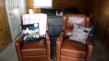 Twee bruine leren fauteuils met gestreepte kussens, een grijze bank en een houten wand met een bord.