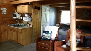 Interior de una cabaña con kitchenette, comedor y zona de estar con un sillón de cuero marrón y un cojín a cuadros.