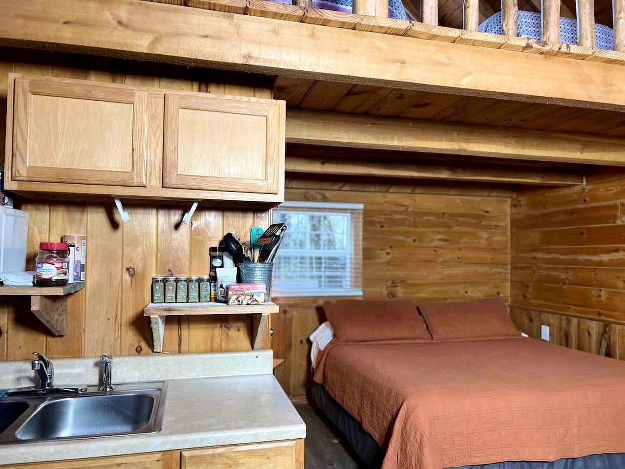 A compact cabin-style room with a kitchenette, a bed with a brown blanket, and wooden walls.