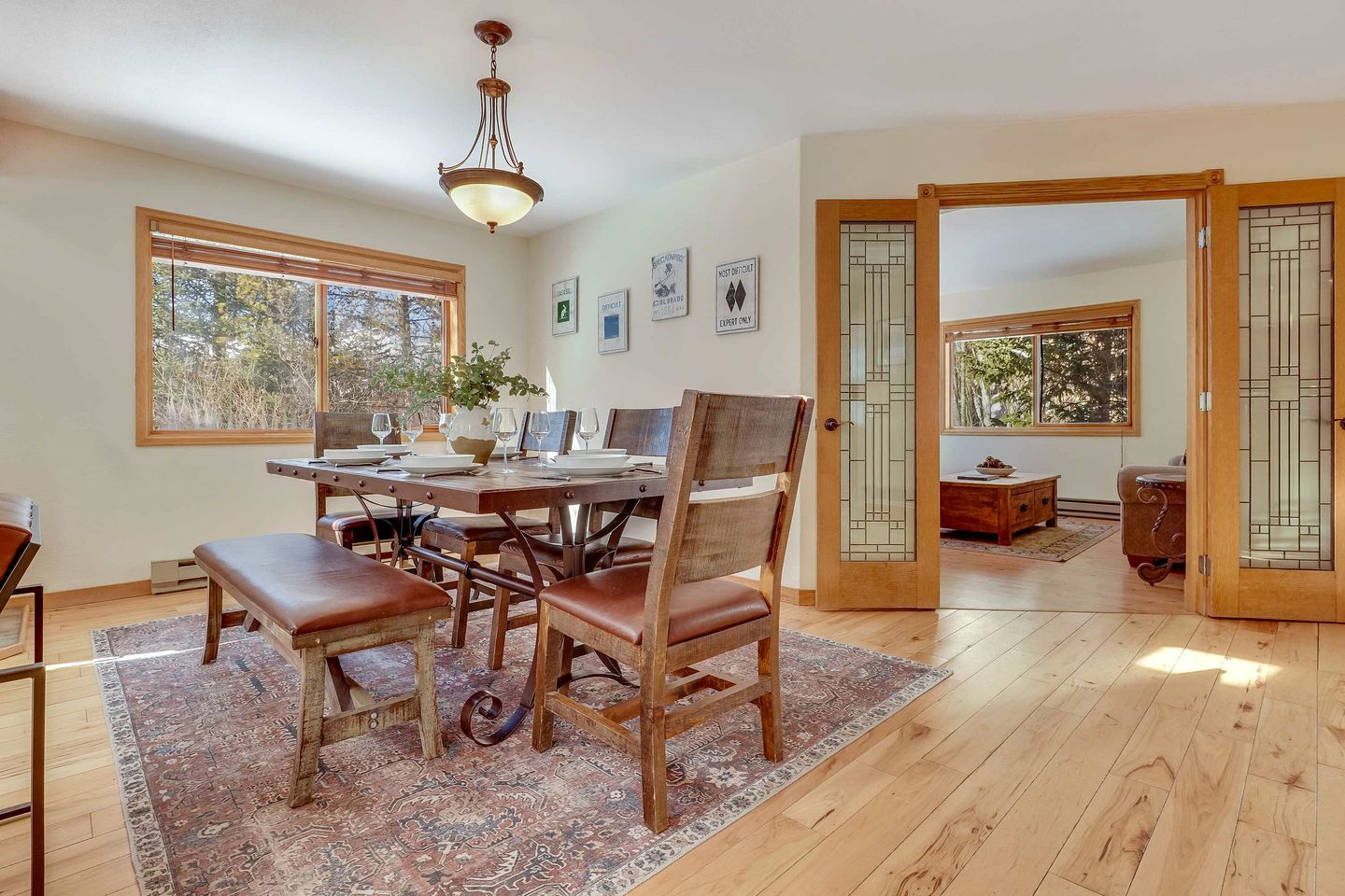 A dining room with a wooden table, chairs, and a chandelier. There are windows with blinds, a rug, and a door with glass panels.