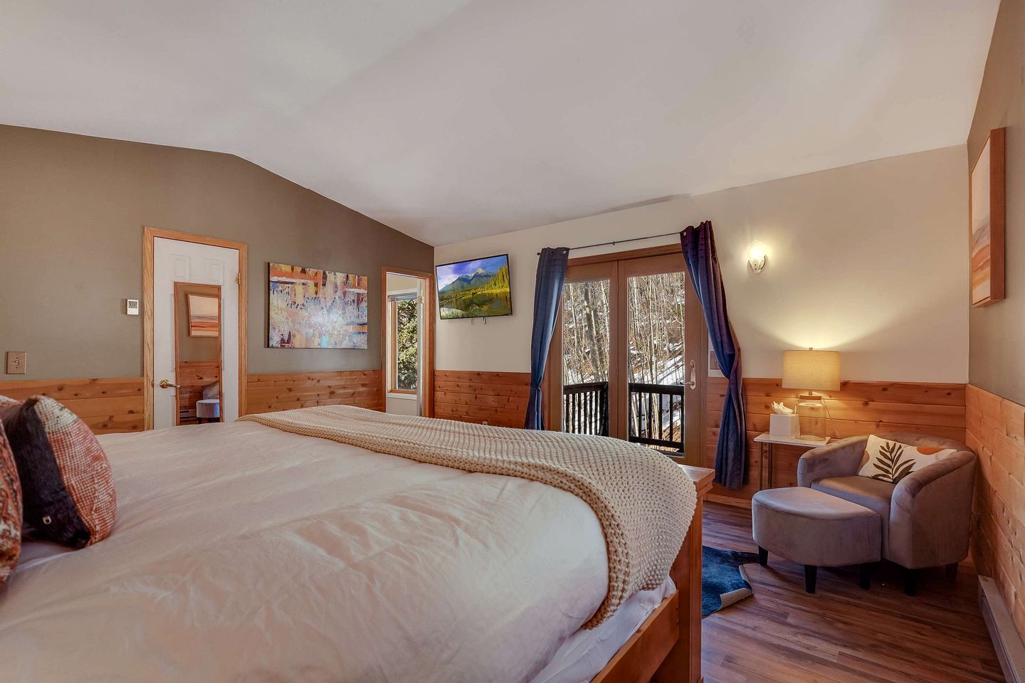 A bedroom with a large bed, a chair, a desk, a painting, and a view of trees through a window.