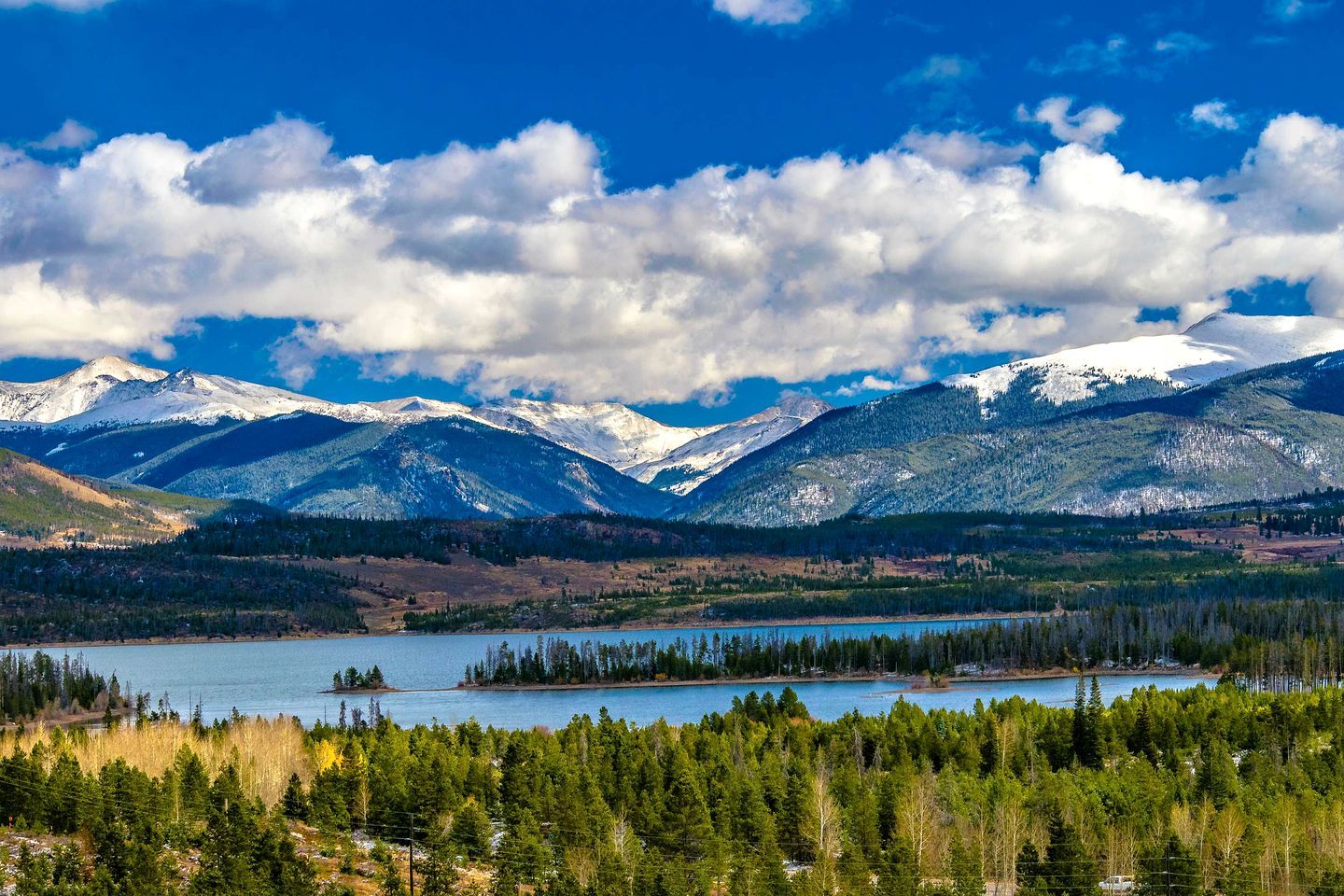 Un paysage montagneux avec un lac, des zones boisées et des sommets enneigés.