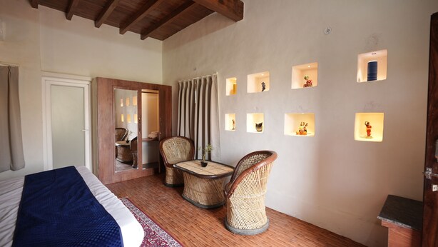 A bedroom with a bed, two wicker chairs, a small table, and a wall with illuminated square cutouts. - Vrinda Tapovan Farmstay (Chhata)