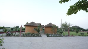 A courtyard with two thatched-roof structures, surrounded by greenery and a paved walkway.