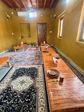 A long dining table with multiple chairs, a wooden ceiling, and a large rug. - Vrinda Tapovan Farmstay (Chhata)