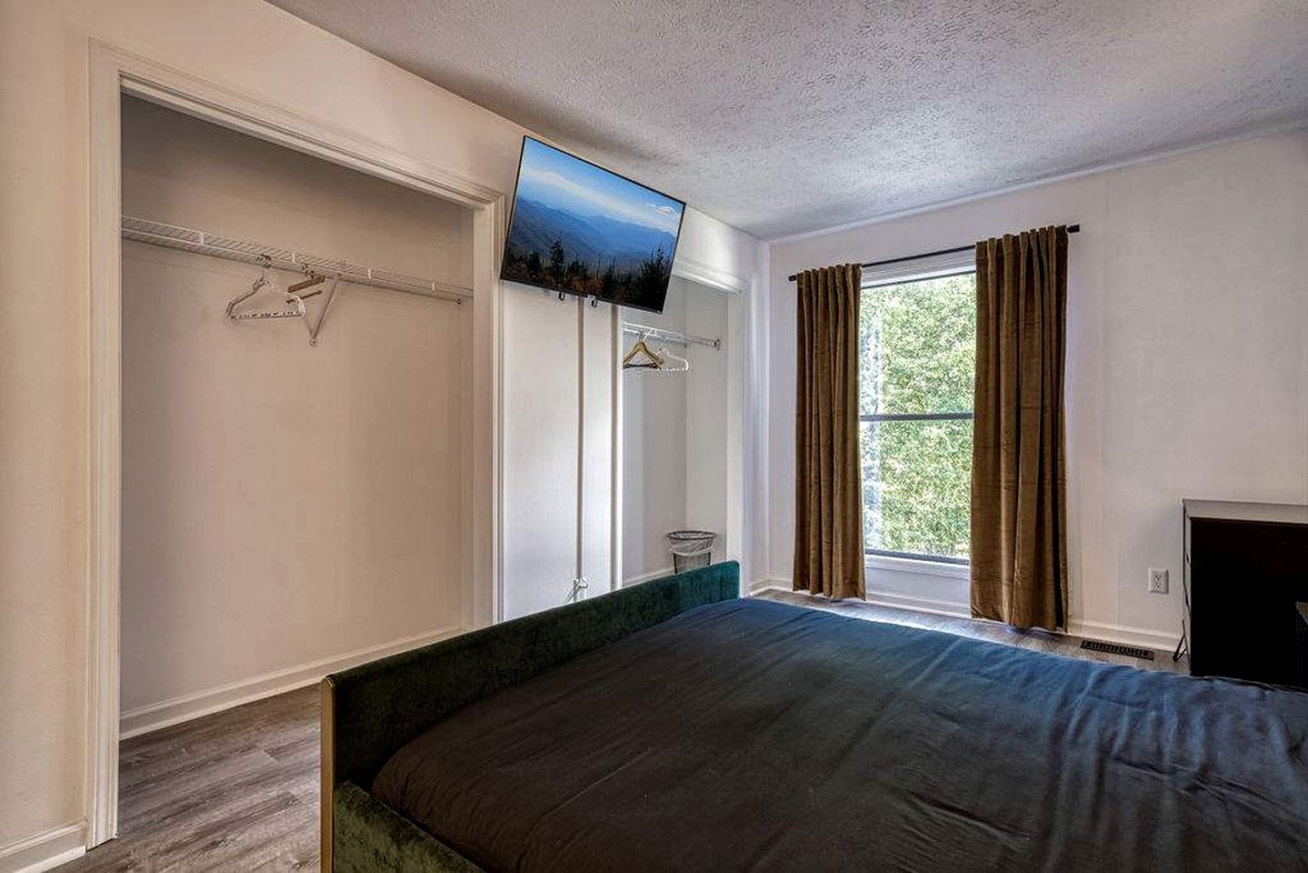 A bedroom with a bed, a television mounted on the wall, a window with curtains, and a closet with hanging clothes.