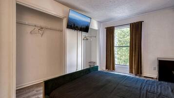 A bedroom with a bed, a television mounted on the wall, a window with curtains, and a closet with hanging clothes.