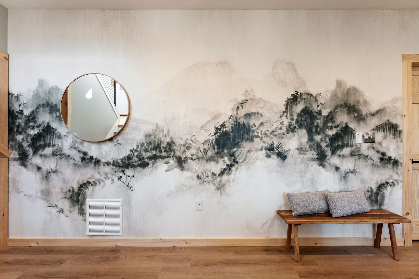 Une pièce avec une fresque murale, un miroir rond, un banc avec des coussins et une porte en bois.