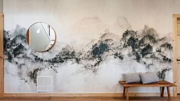 Une pièce avec une fresque murale, un miroir rond, un banc avec des coussins et une porte en bois.