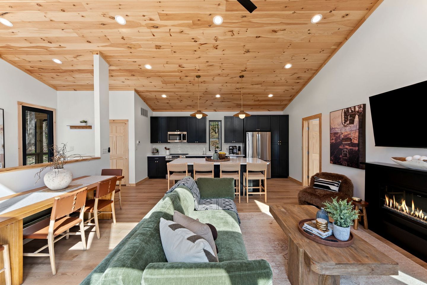 A modern living room with a green sofa, wooden dining table, and chairs, a kitchen with dark cabinets and stainless steel appliances, and a fireplace.