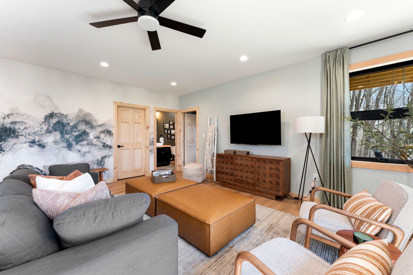 A modern living room with a sofa, ottoman, and armchairs, a flat-screen TV, a ceiling fan, and a large window with a view of trees.