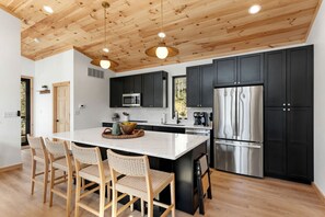 Une cuisine moderne avec un plafond en bois, des meubles de couleur noire, des appareils électroménagers en acier inoxydable et un îlot central doté de chaises.