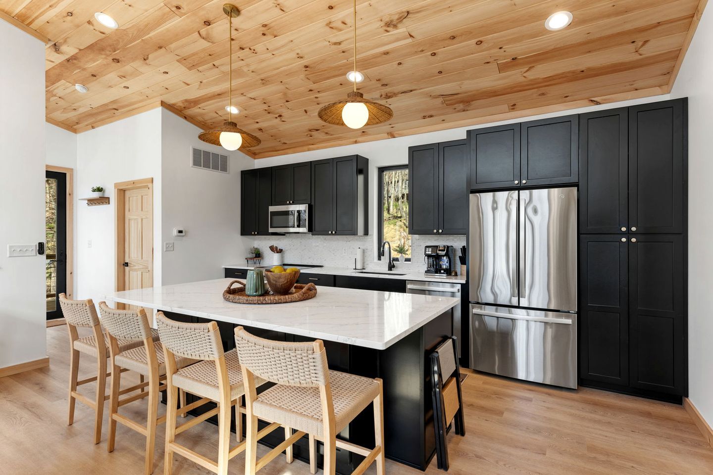 A modern kitchen with a wooden ceiling, black cabinets, stainless steel appliances, and a central island with chairs.