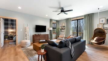 Ein modernes Wohnzimmer mit einem dunklen Sofa, einem Fernseher, einem Deckenventilator und einem großen Fenster mit Vorhängen.