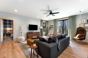 A modern living room with a dark sofa, a television, a ceiling fan, and a large window with curtains.