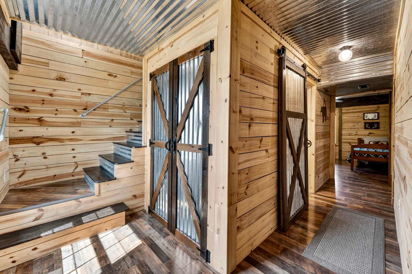 A wooden interior with a staircase, a metal-framed door, and a bed in the background.