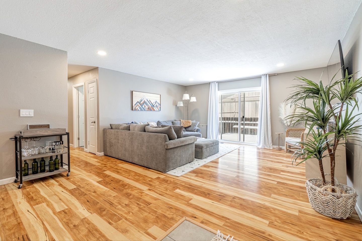 Un salon moderne avec un canapé gris, un plancher en bois, une table basse en verre et une grande fenêtre avec des rideaux.