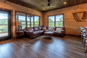 A cabin interior with a wooden ceiling, large windows, a leather sofa, a round coffee table, and a dining area with chairs. - Spacious Luxury Cabin with Game Room near Pigeon Forge Attractions – Gatlinburg, TN (Gatlinburg)